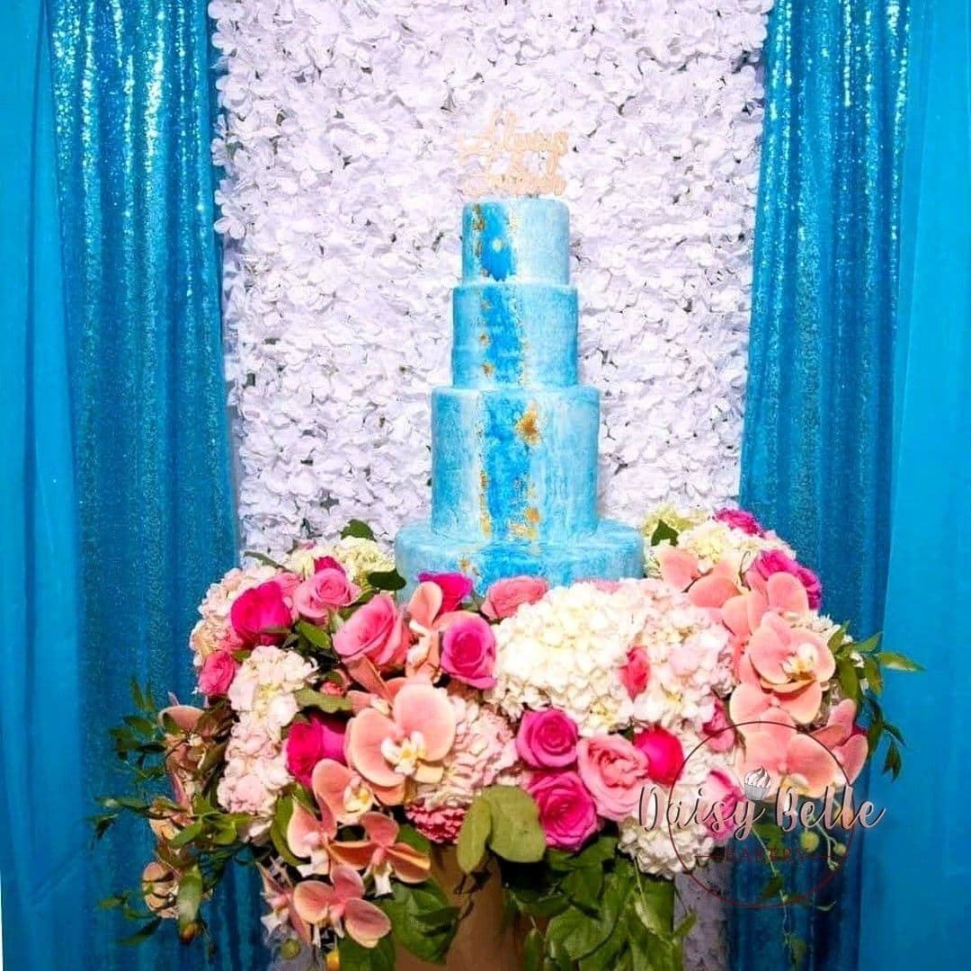 A four-tier wedding cake painted in watercolour blues with gold leaf, standing against a white flower wall between teal drapes, banked with pink roses, orchids and white hydrangeas.