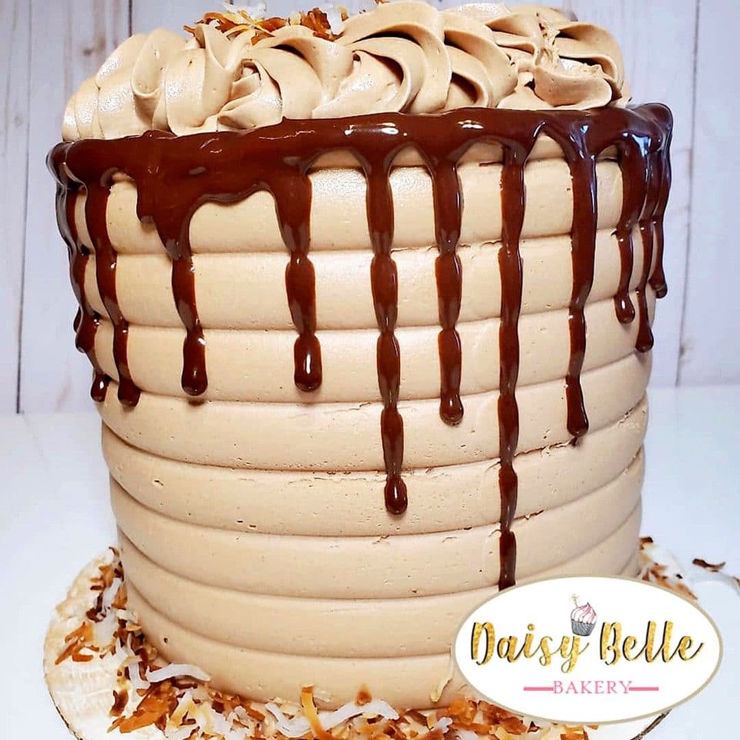 A tall mocha buttercream cake finished in horizontal ridges, with dark chocolate ganache running down the sides, piped swirls across the top and toasted coconut scattered around the base.
