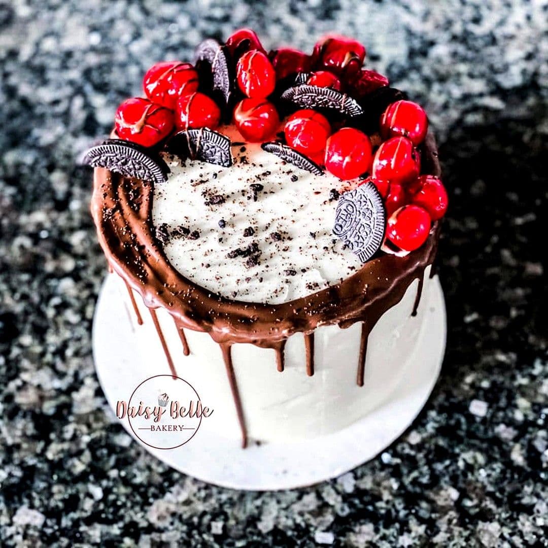 A tall white buttercream cake under a chocolate drip, topped with a disc of crushed-cookie cream and ringed with glossy red cherries and halved sandwich cookies.