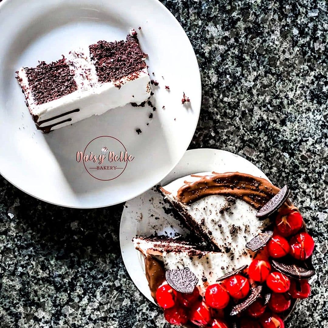 An overhead view of a cut cookies-and-cream cake on a speckled granite counter, its edge crowded with glossy red cherries and chocolate sandwich cookies, with one slice lifted onto a white plate beside it.
